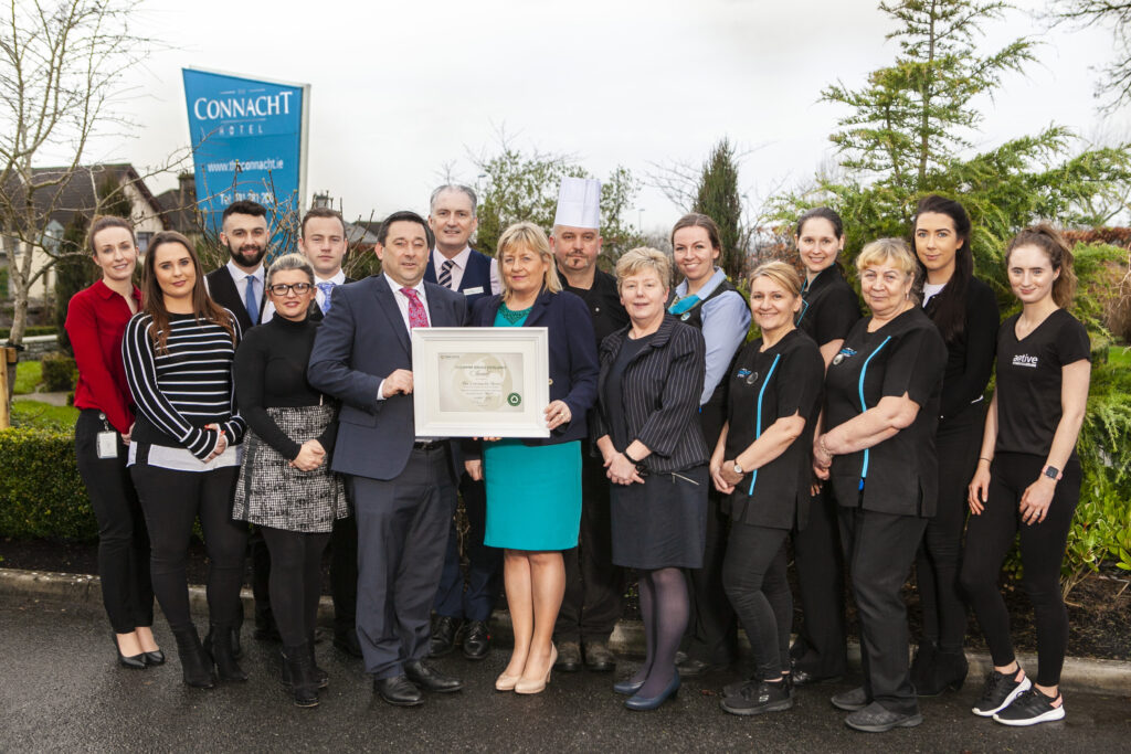 Hotel Team Holding an Award Outdoors – A large team of hospitality professionals, including chefs, front-desk staff, and management, pose outdoors in front of a Connacht Hotel banner, proudly holding a framed certificate. They are dressed in business and work attire