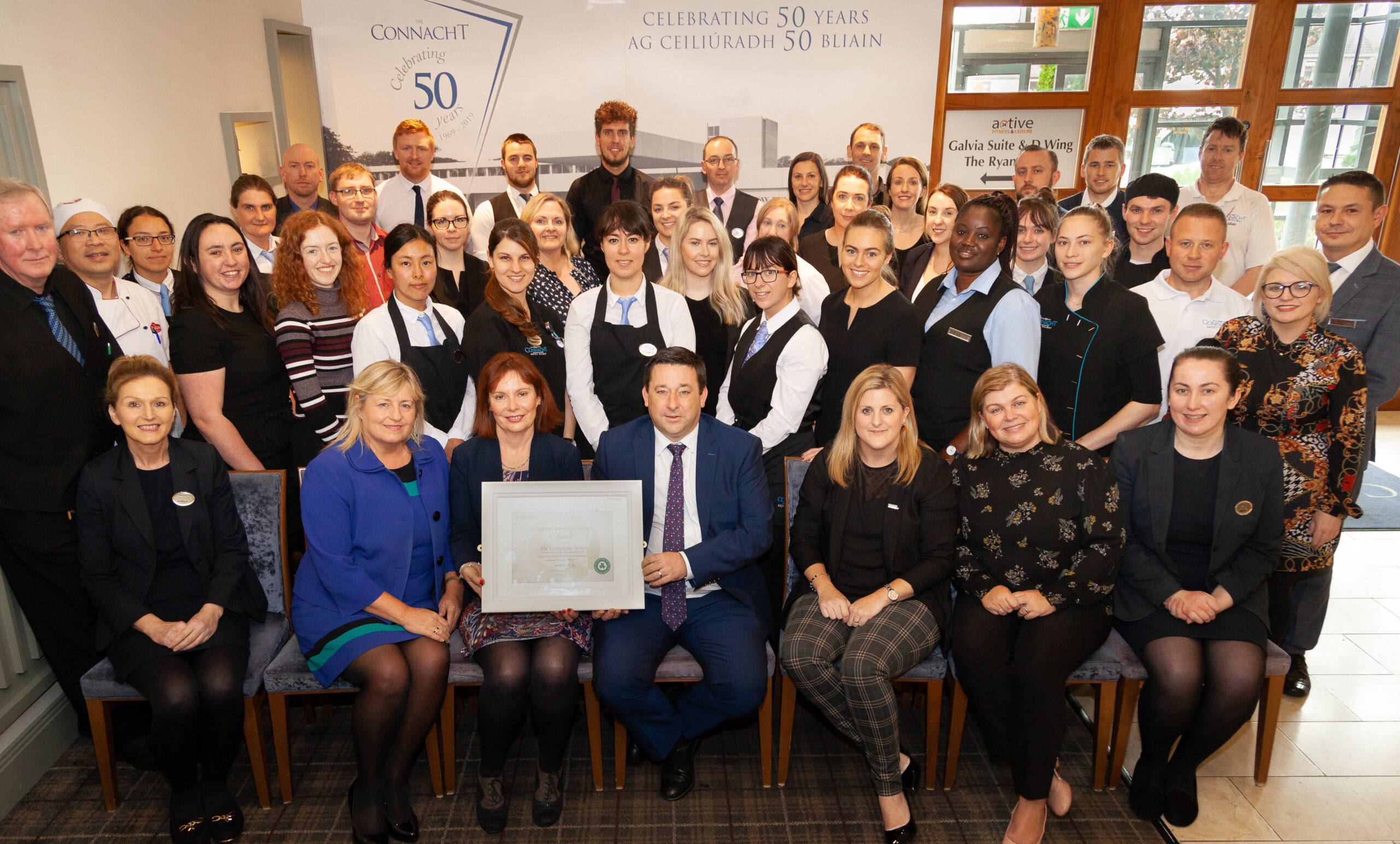 Large Group of Hospitality Staff Holding an Award Indoors – A team of hotel staff and management, dressed in business attire and uniforms, gather indoors to celebrate an award. A banner in the background indicates the hotel's 50-year celebration.