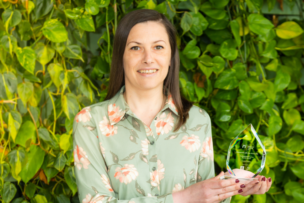 Woman Holding an Award in Front of a Green Wall – A woman in a floral-patterned green blouse smiles while holding a glass award. The lush green plant wall behind her enhances the fresh and celebratory feel of the image.