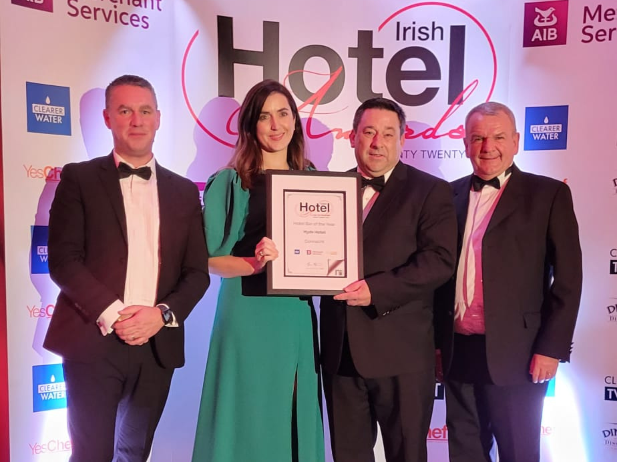 Four People Holding an Award at the Irish Hotel Awards – A group of four well-dressed individuals, including a woman in a green dress and men in suits, hold a framed award certificate at the Irish Hotel Awards. The event branding and sponsors appear in the background.