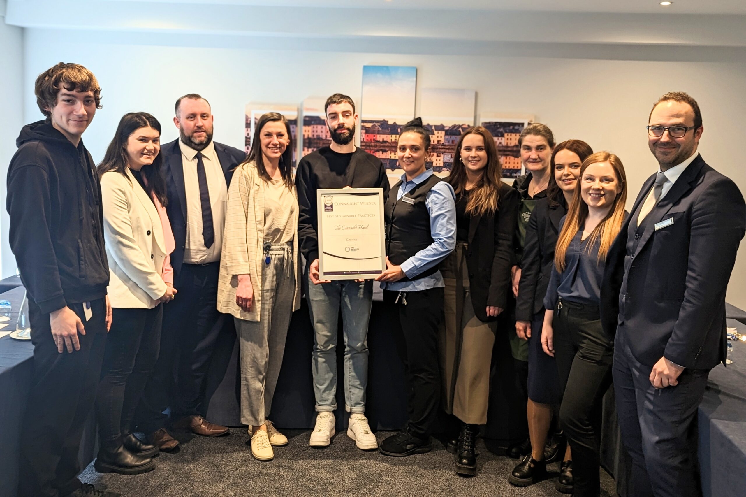 Group of Employees Holding an Award in a Conference Room – A diverse group of employees, including management and front-line staff, pose in a professional setting, holding a framed award. A scenic Galway cityscape print is visible in the background.