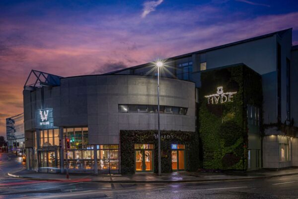 A contemporary, stylish building illuminated at twilight, featuring the "HYDE" and "WYLDE" signage. The exterior showcases a combination of sleek concrete architecture and a lush green wall. Large glass windows reveal a warmly lit interior, while a streetlight casts a glow on the sidewalk, enhancing the inviting ambiance of the location.