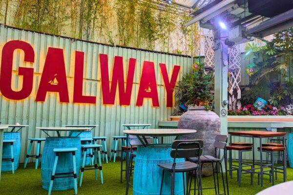 The beer garden at An Púcán - A lively and colorful outdoor bar area featuring bold red "GALWAY" lettering on a light green corrugated metal wall. The space is decorated with greenery, hanging plants, and bright blue barrel-style tables with matching stools. The artificial grass flooring and eclectic decor create a fun and relaxed atmosphere, enhanced by overhead lighting and decorative elements.