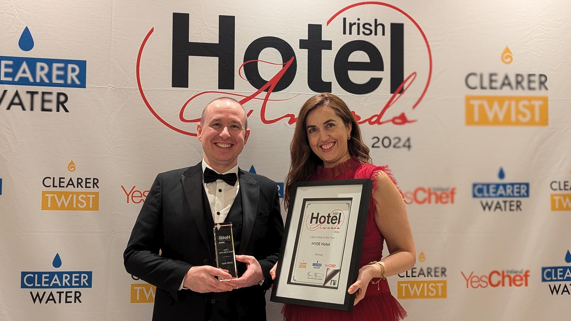 Two People Holding an Award at the Irish Hotel Awards 2024 – A man in a tuxedo and a woman in a red dress stand smiling at the Irish Hotel Awards 2024, holding a framed certificate and a trophy. The backdrop features event sponsors and branding.