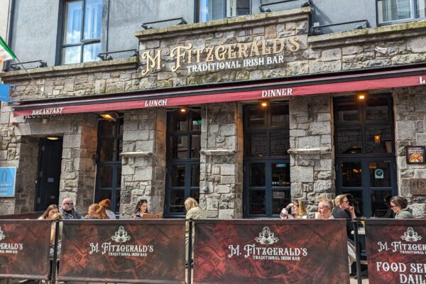 M. Fitzgerald’s Traditional Irish Bar – A lively Irish pub with a rustic stone exterior, outdoor seating, and red awnings advertising breakfast, lunch, and dinner service.