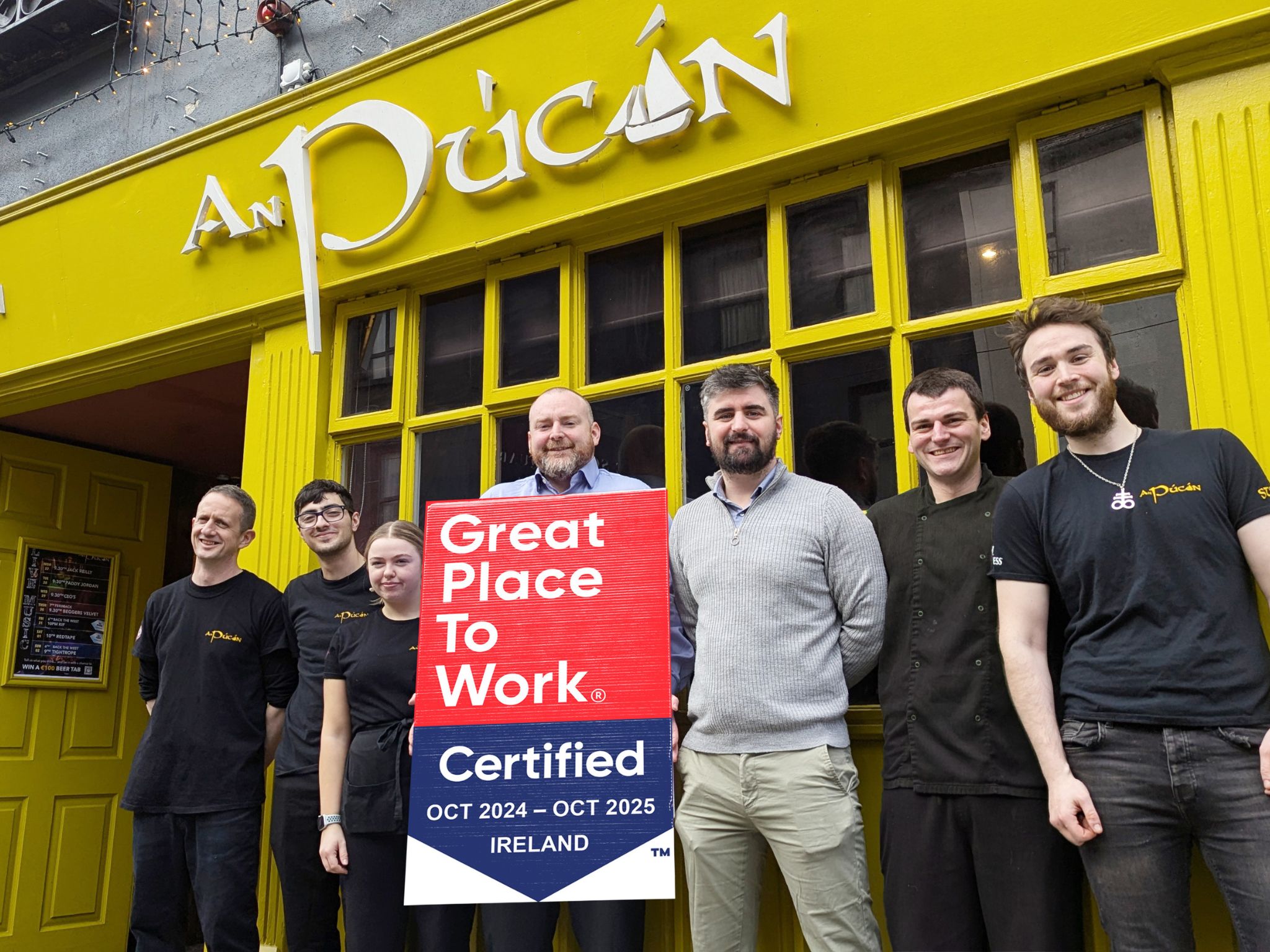 An Púcán Team Holding Great Place to Work Certification – The team of An Púcán, a vibrant pub with a bright yellow exterior, poses cheerfully outside while holding a "Great Place to Work" certification sign for October 2024 - October 2025.