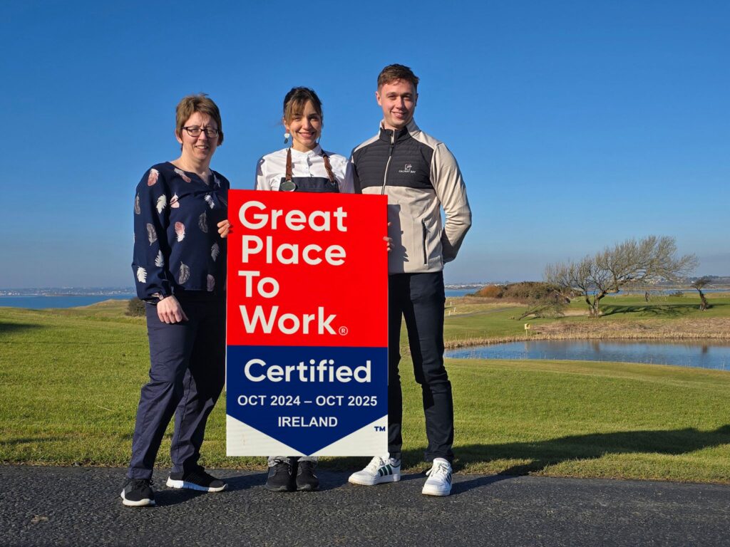 Three employees pose on a scenic golf course with rolling green hills and a calm body of water in the background. They hold a "Great Place to Work Certified" sign for October 2024 to October 2025 in Ireland. The team members wear a mix of smart casual and golf attire, with one in a chef's uniform. The clear blue sky and open landscape create a picturesque setting for the recognition.