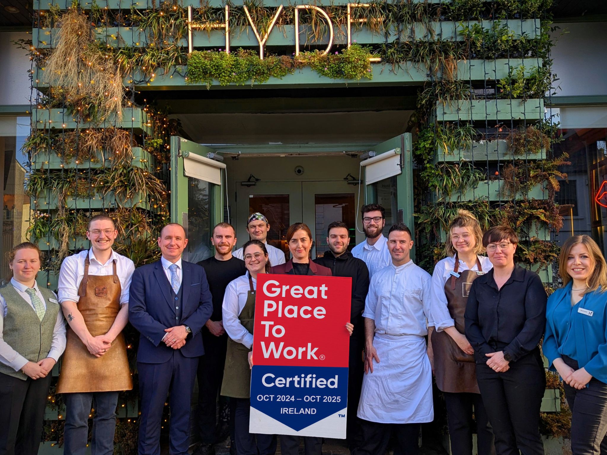 A diverse team of hospitality professionals, including chefs, servers, and management staff, stands outside Hyde Bar, a trendy establishment with lush greenery decorating its exterior. They hold a "Great Place to Work Certified" sign for October 2024 to October 2025 in Ireland. The team wears a mix of stylish uniforms, suits, and casual business attire, smiling as they celebrate their achievement.