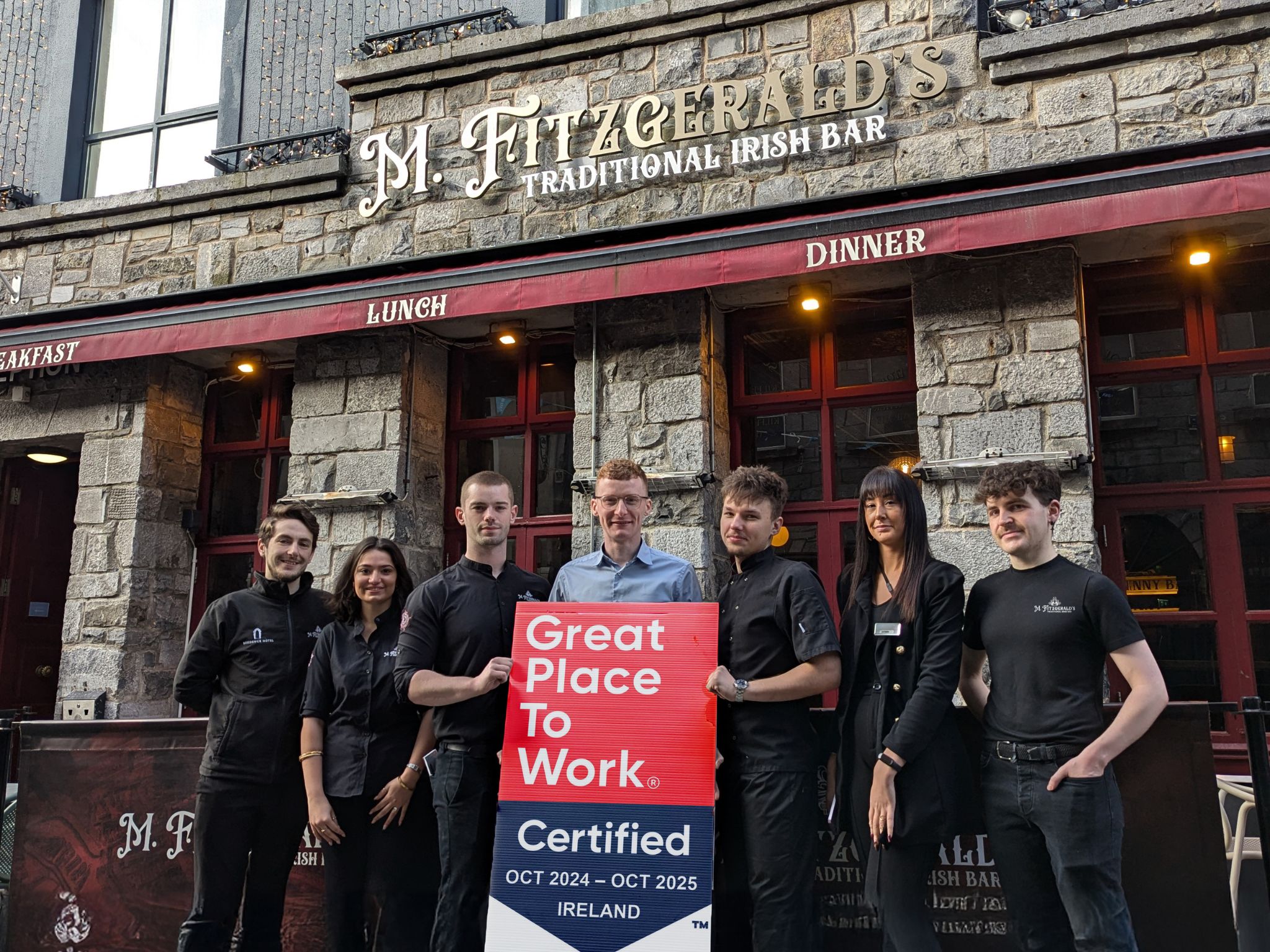 A group of seven staff members stands outside M. Fitzgerald's Traditional Irish Bar, a rustic stone-fronted establishment with red-framed windows and signage for breakfast, lunch, and dinner. They hold a "Great Place to Work Certified" sign for October 2024 to October 2025 in Ireland. The team members wear a mix of black uniforms and casual attire, smiling in front of the Irish pub.