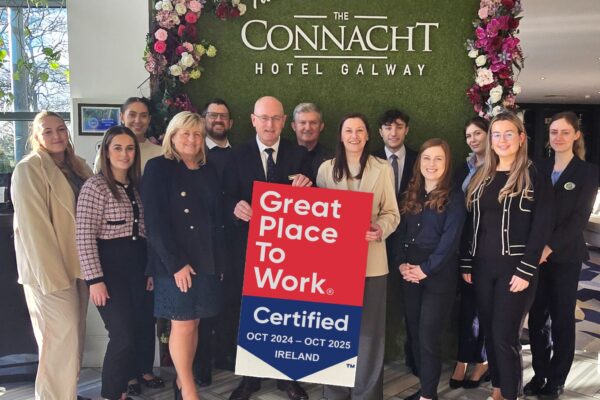 The Connacht Hotel Team Celebrating Great Place to Work Certification – A group of professionally dressed staff members from The Connacht Hotel Galway posing in front of a lush green wall with the hotel's name and floral decorations. They proudly hold a "Great Place to Work" certification sign for October 2024 - October 2025.