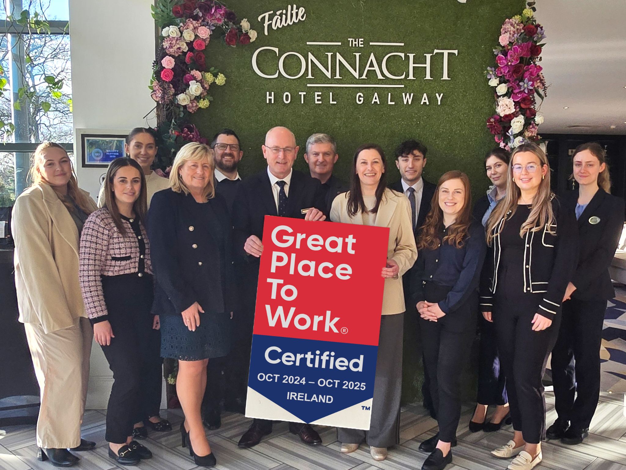 A large group of professionally dressed staff members gathers inside the lobby of The Connacht Hotel Galway. They hold a "Great Place to Work Certified" sign for October 2024 to October 2025 in Ireland. Behind them is a green wall with the hotel's name in white lettering, decorated with floral arrangements. The team members are dressed in formal attire, including suits, blazers, and smart casual outfits, all smiling at the camera.