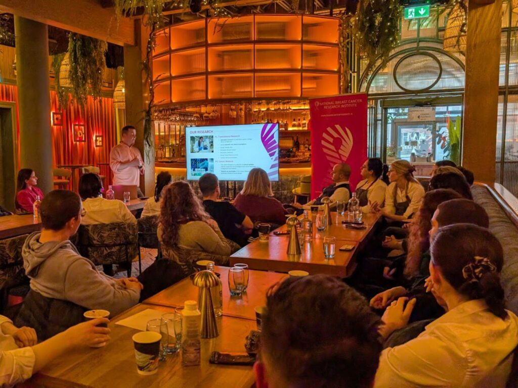 Breast Cancer Awareness Presentation – A group of people seated in a warmly lit venue listen attentively to a speaker giving a presentation on breast cancer awareness, with a National Breast Cancer Research Institute banner in the background.