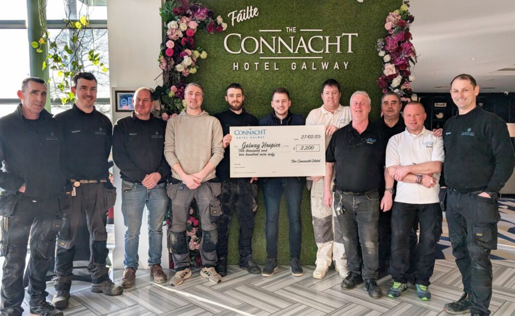 Connacht Hotel Galway Charity Donation – A group of staff members and workers from The Connacht Hotel Galway stand together holding a large charity cheque for Galway Hospice, posing in front of a floral wall with the hotel's name.